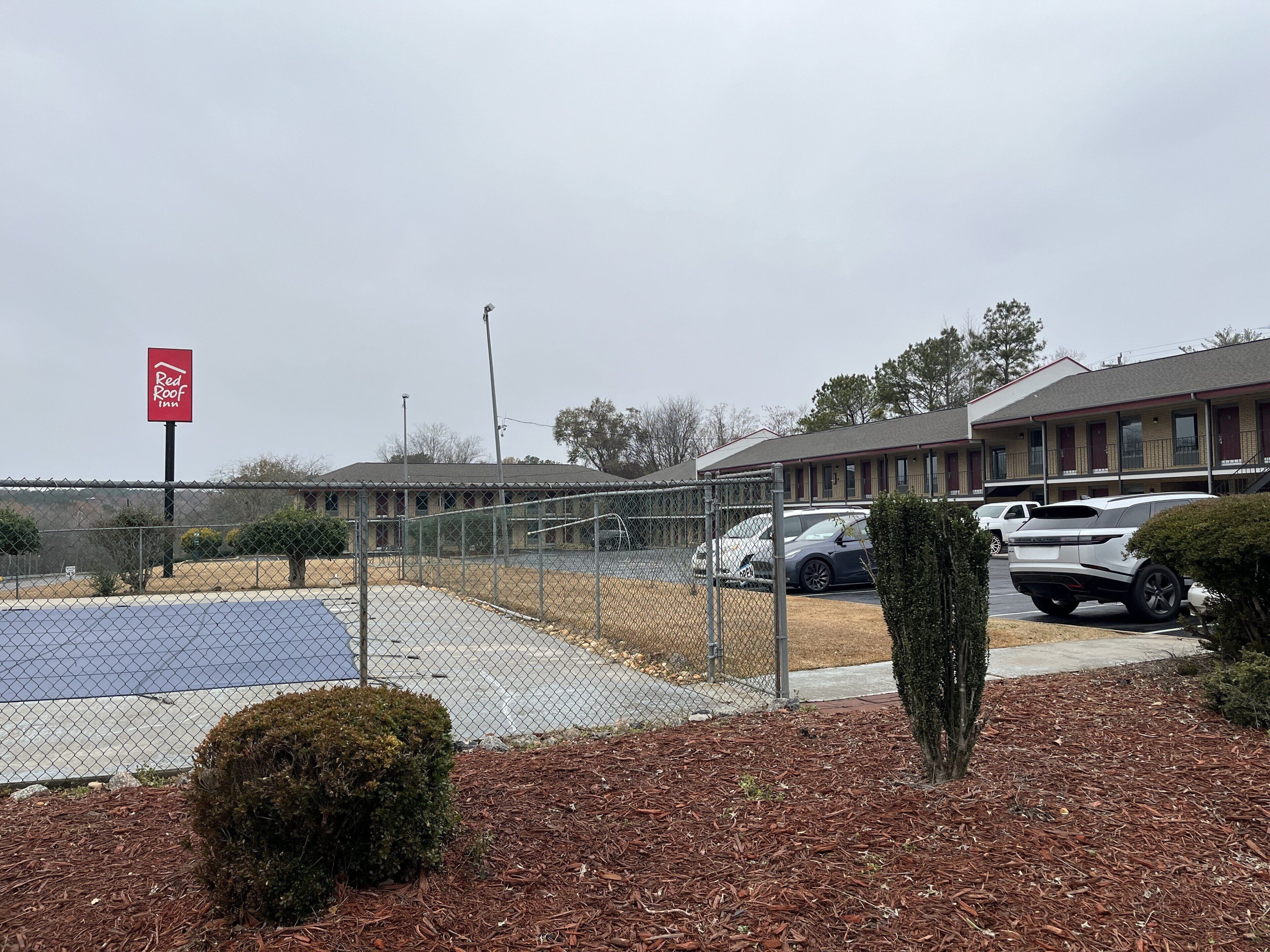 Ein durch Zäune abgegrenzter Parkbereich mit einem Red Roof Inn Schild, ein Gebäude mit rotem Dach und einem wolkenlosen Himmel.