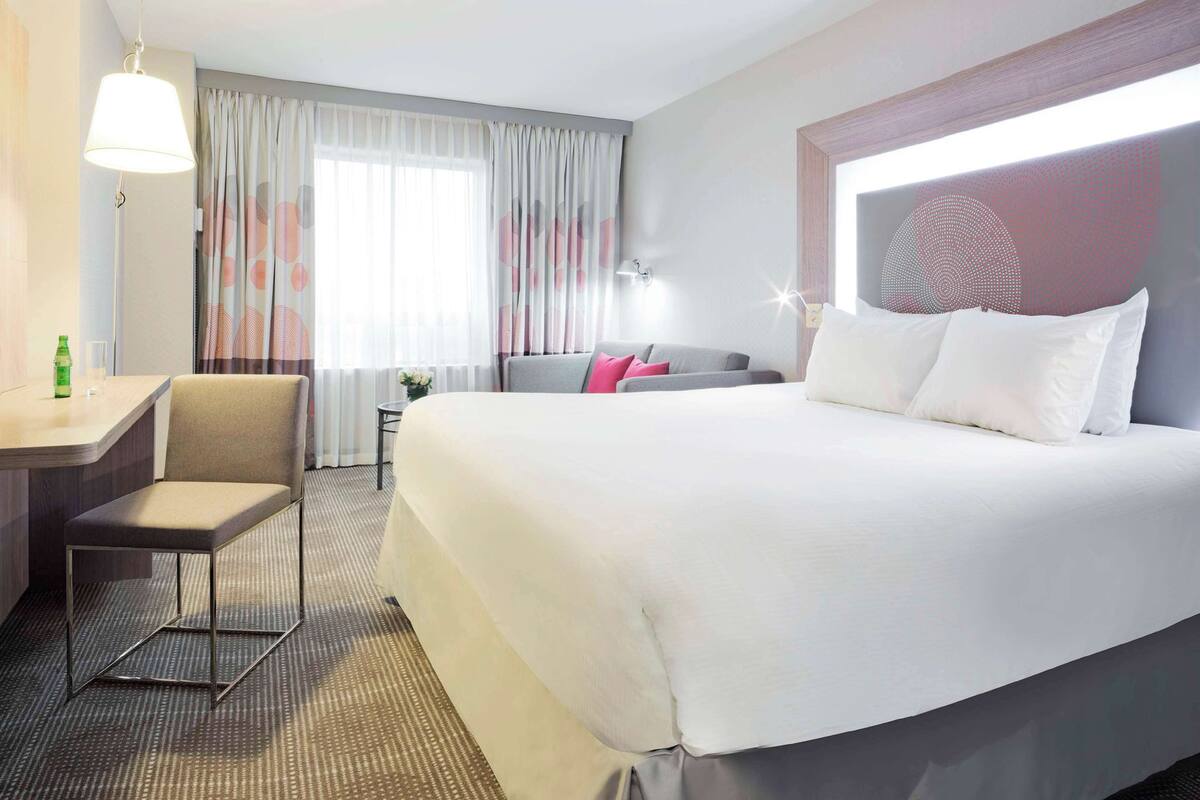Habitación de hotel con una cama grande, un escritorio con silla, una mesita con una botella y un vaso, y una ventana con cortinas.