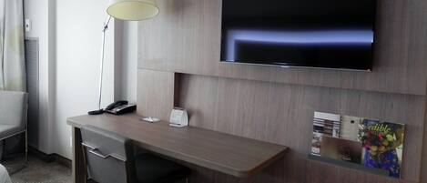 A hotel room with a desk, a chair, a TV mounted on the wall, a lamp, and a small bulletin board with a poster.