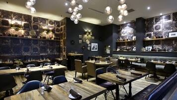 Um restaurante com mesas e cadeiras de madeira, um espaço de bar e uma parede com quadros e um relógio.