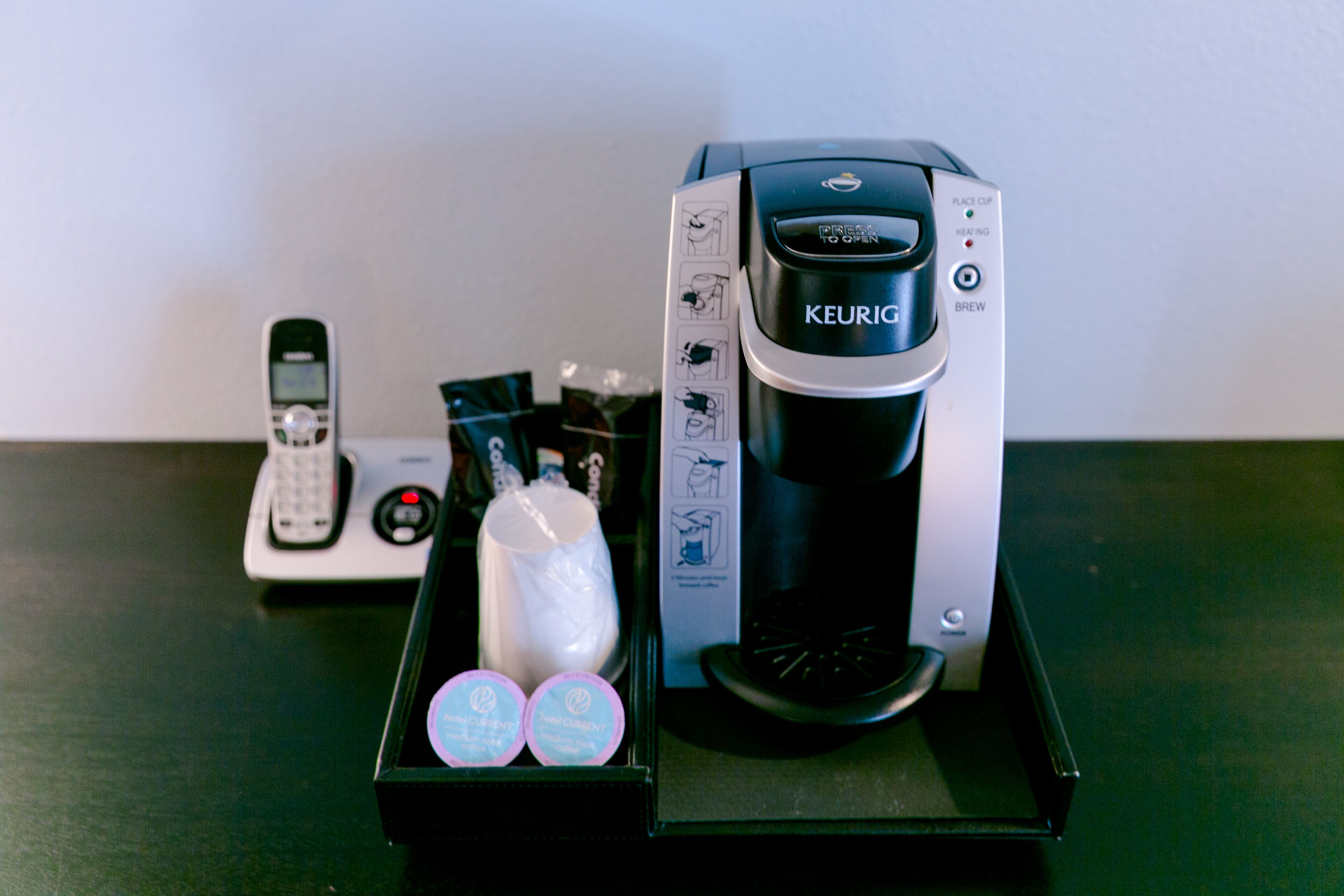 A Keurig coffee maker with a drip tray, a remote control, and two Keurig pods on a table.
