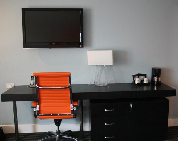 A hotel room with a black desk, an orange chair, a TV mounted on the wall, a white lamp, and a coffee maker.