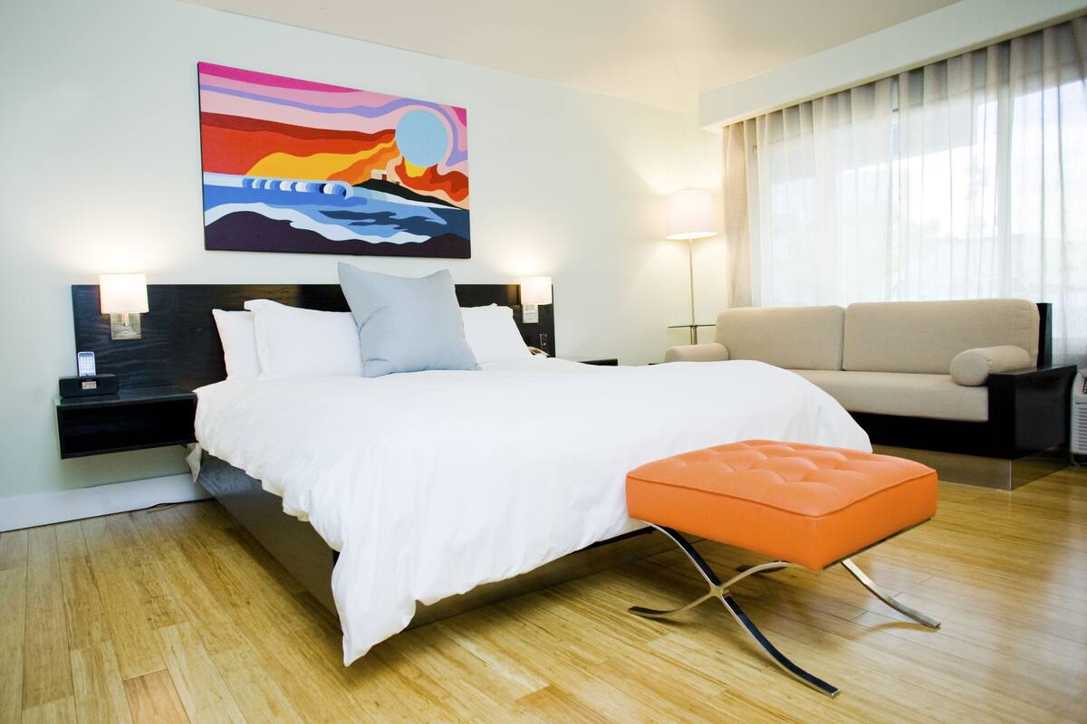Quarto de hotel moderno com uma cama grande, um banquinho laranja, um sofá, um quadro na parede e uma mesa de cabeceira com uma luminária.