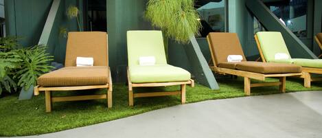 Trois fauteuils de jardin en bois, dotés de coussins, sont placés sur une surface en béton, entourés de gazon artificiel vert et d’un bâtiment à l’architecture moderne.