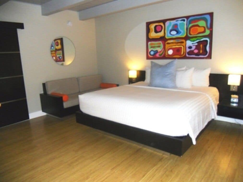 Une chambre d’hôtel moderne avec un grand lit, une table de chevet, une commode, une œuvre d’art accrochée au mur et un miroir rond.