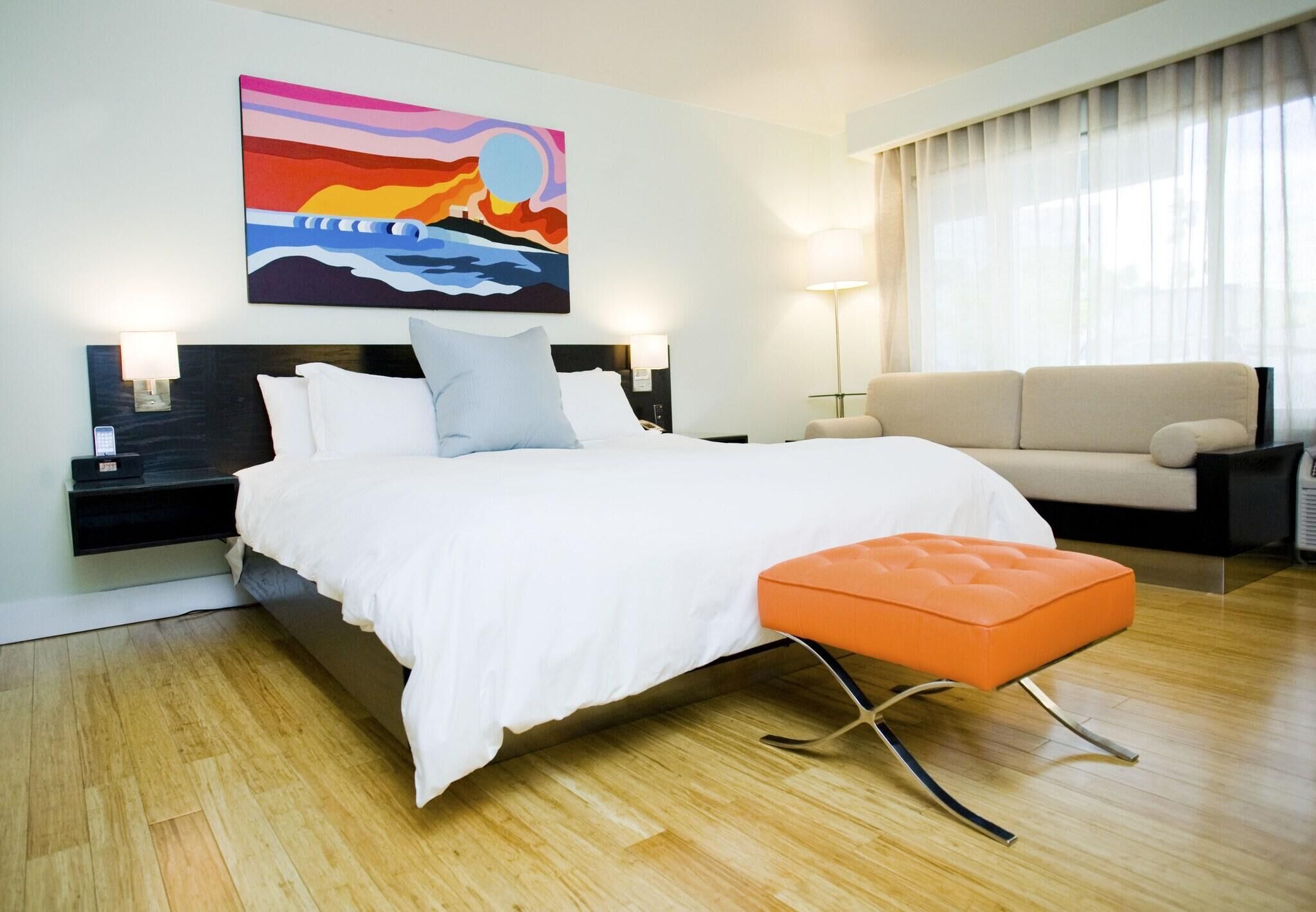 A modern hotel room with a large bed, a sofa, a painting on the wall, and a nightstand with a lamp.