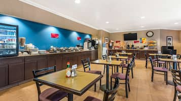 A hotel lobby with a self-service beverage station, multiple tables and chairs, a clock, and a television on the wall.