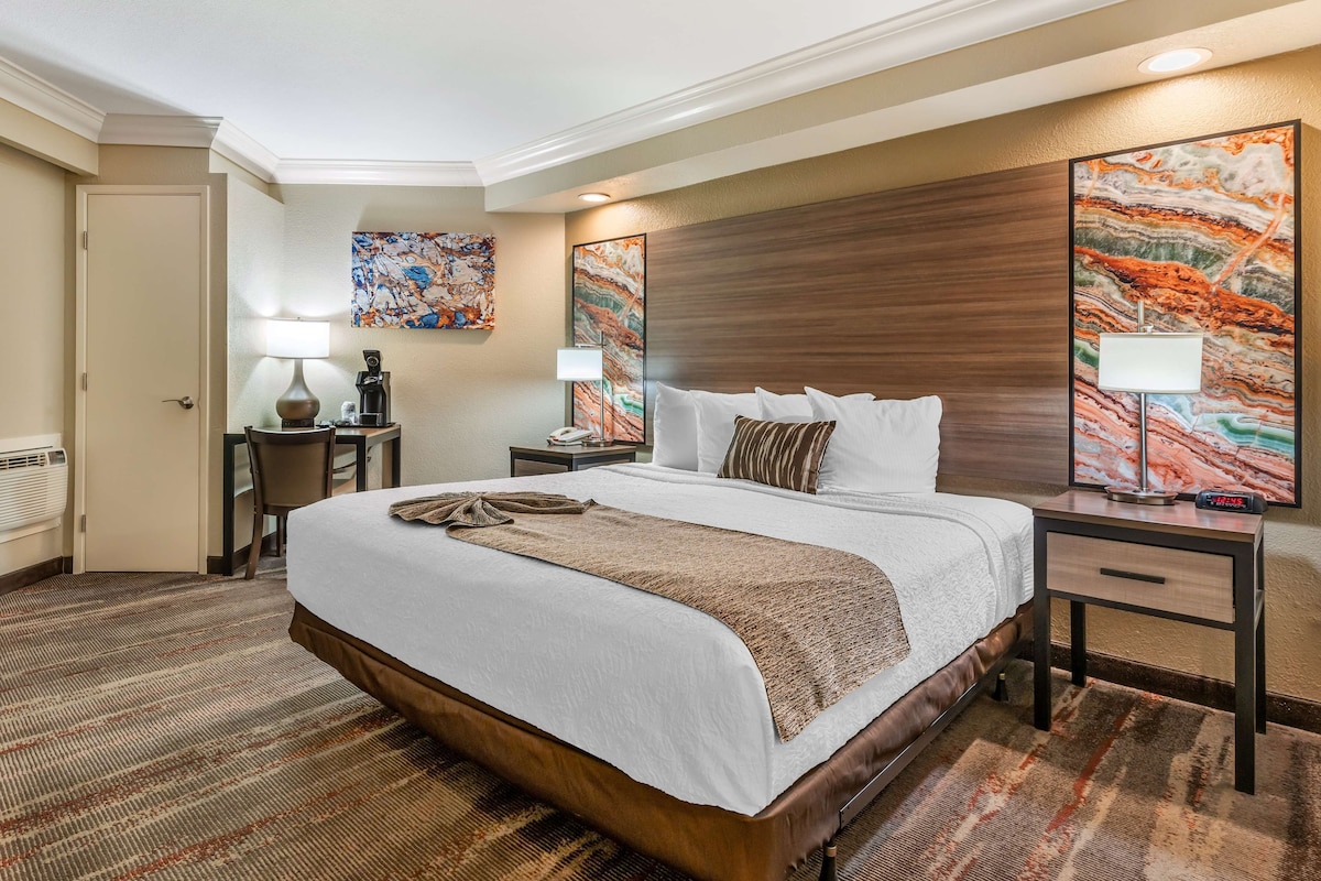 Une chambre d’hôtel avec un grand lit, deux tables de chevet, un bureau et une chaise. Il y a deux lampes fixées au mur et un tableau au-dessus du lit.