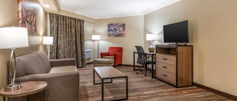 Habitación de hotel con sofá, una mesita de centro, escritorio con silla, televisor sobre un mueble bajo y un ventanal con cortinas.