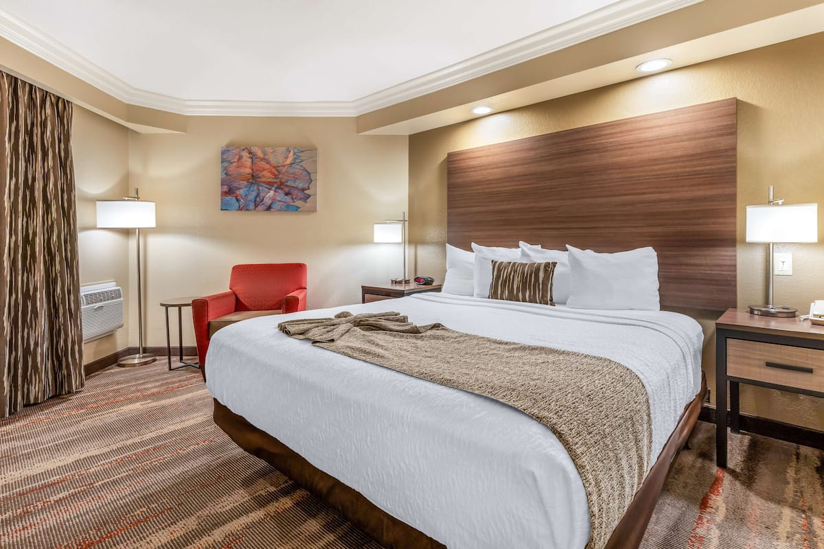A hotel room with a large bed, a red armchair, a small table, and a painting on the wall.