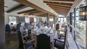 A spacious dining area with round tables set for a meal, wooden beams on the ceiling, and large windows offering a view of the outdoors. - Rancho Viejo Resort and Country Club (Olmito)