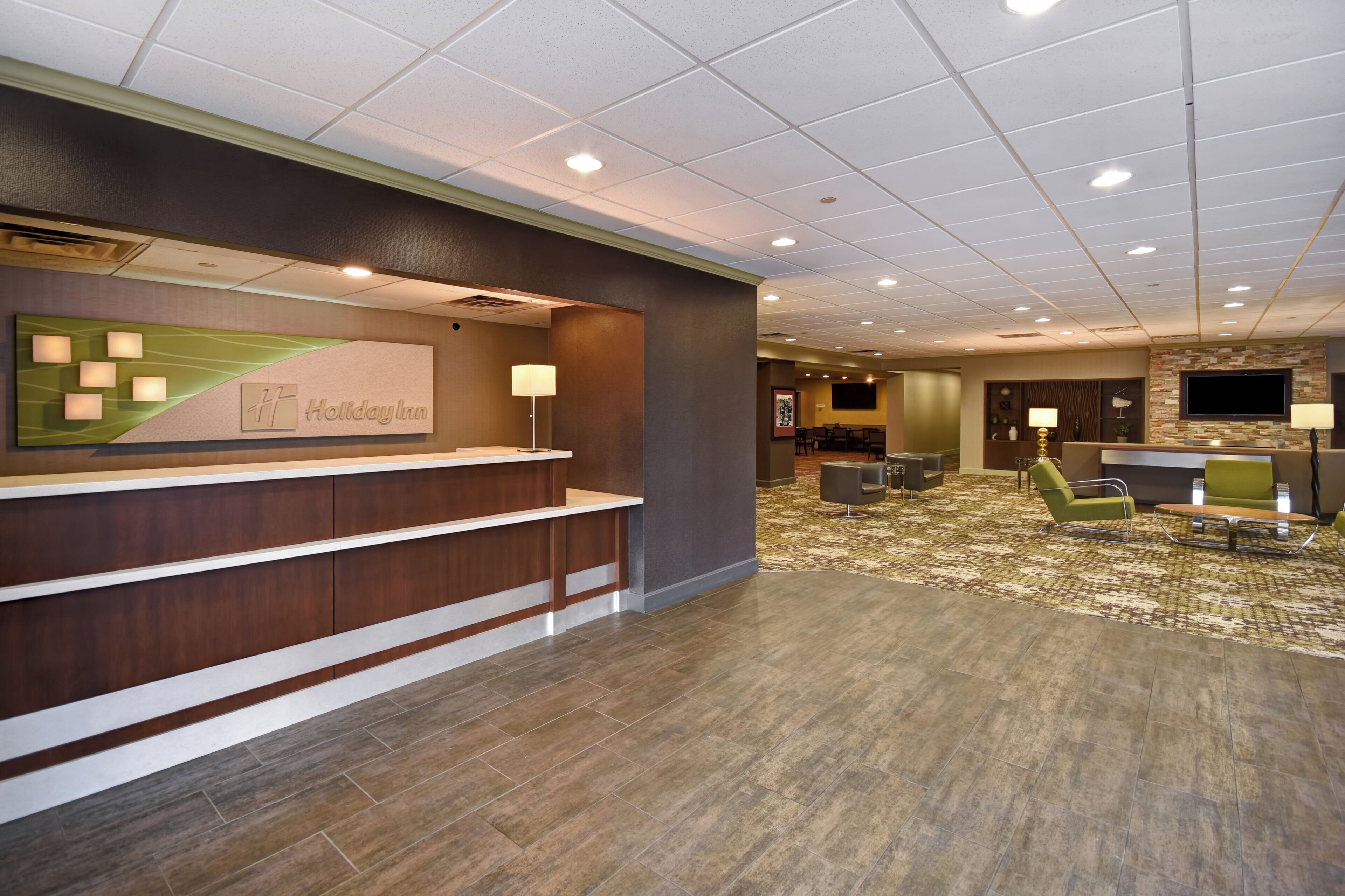 A hotel reception area with a reception desk, a wall-mounted TV, and a seating area with chairs and a table.