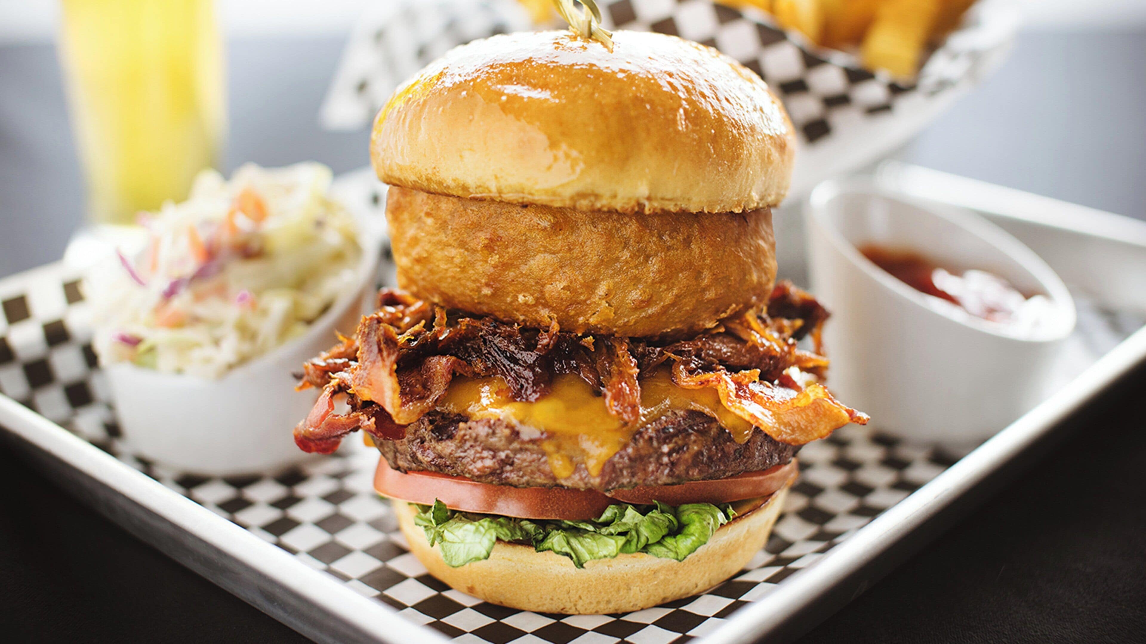 A burger with bacon, cheese, and lettuce on a bun, served on a checkered tray with a side of coleslaw and a dipping sauce.