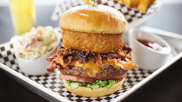 A burger with bacon, cheese, and lettuce on a bun, served on a checkered tray with a side of coleslaw and a dipping sauce.