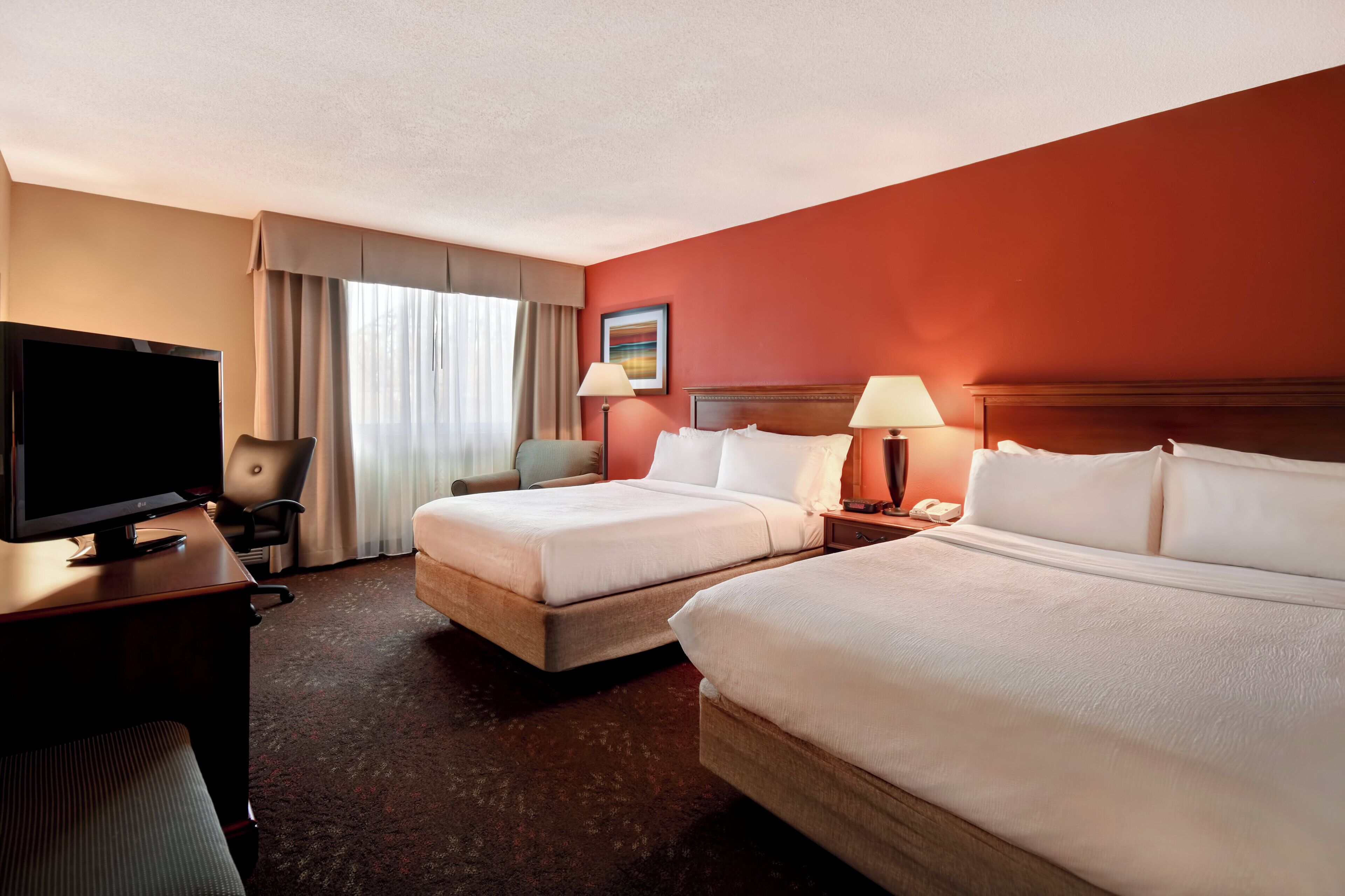 Ein Hotelzimmer mit zwei Betten, einem Schreibtisch mit Fernseher, einem Sessel, einer Lampe und einem Bild an der Wand.