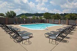 A swimming pool with lounge chairs and tables arranged around it, a fenced area, and a view of trees and buildings in the background.