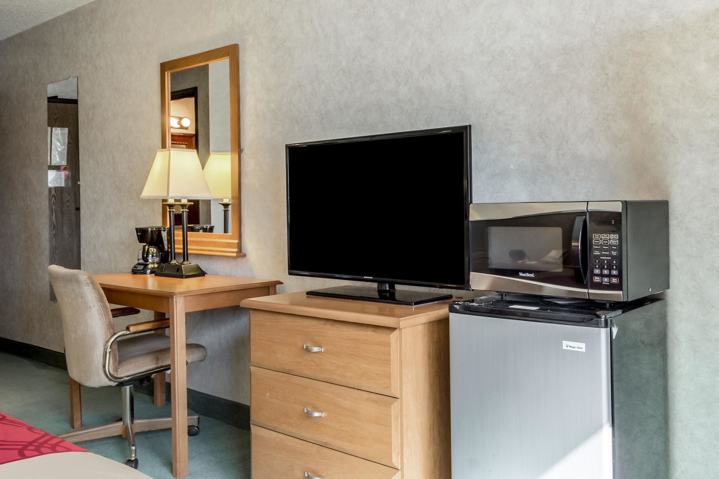 Une chambre d’hôtel équipée d’un bureau en bois, d’une télévision, d’un four à micro-ondes, d’un réfrigérateur, d’une chaise et d’un miroir.