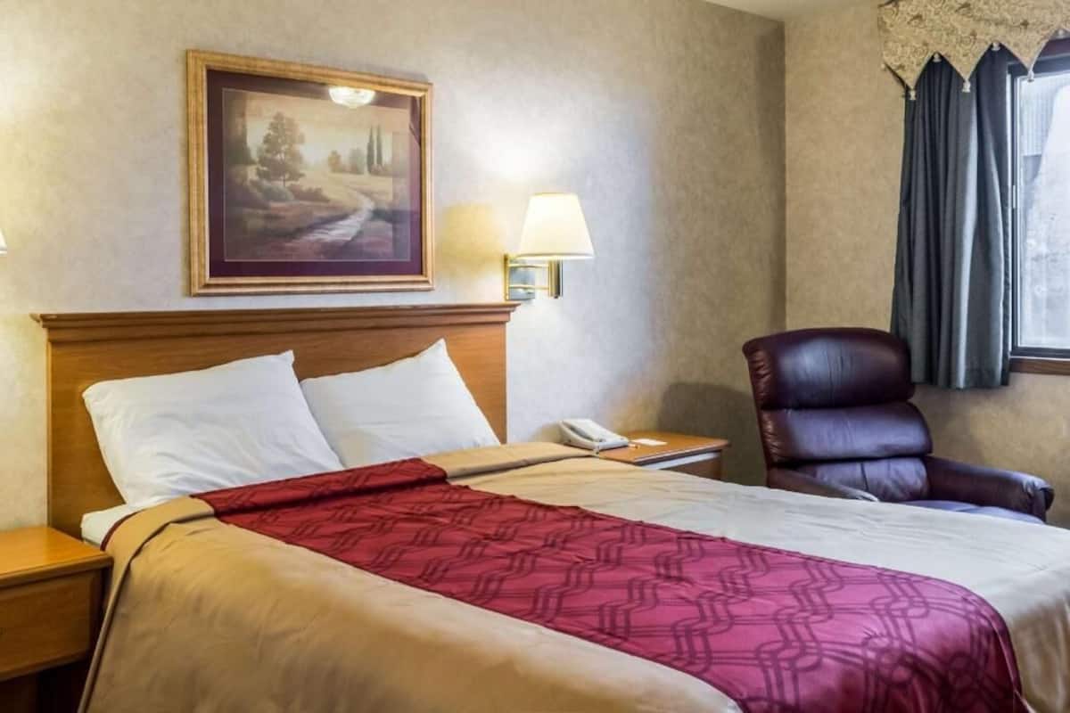 A hotel room with a bed, a nightstand, a telephone, a leather chair, a painting on the wall, and a window with curtains.