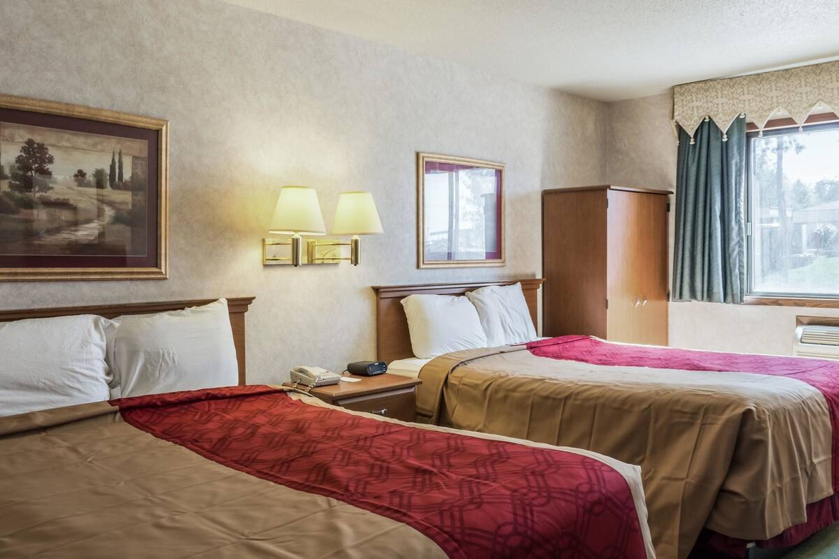 A hotel room with two beds, a nightstand, a lamp, a painting, and a window with curtains.
