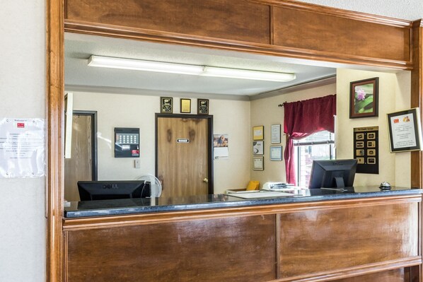 Un área de recepción con un escritorio de madera, una computadora y un teléfono. Hay diplomas enmarcados en la pared y una ventana con cortina roja.