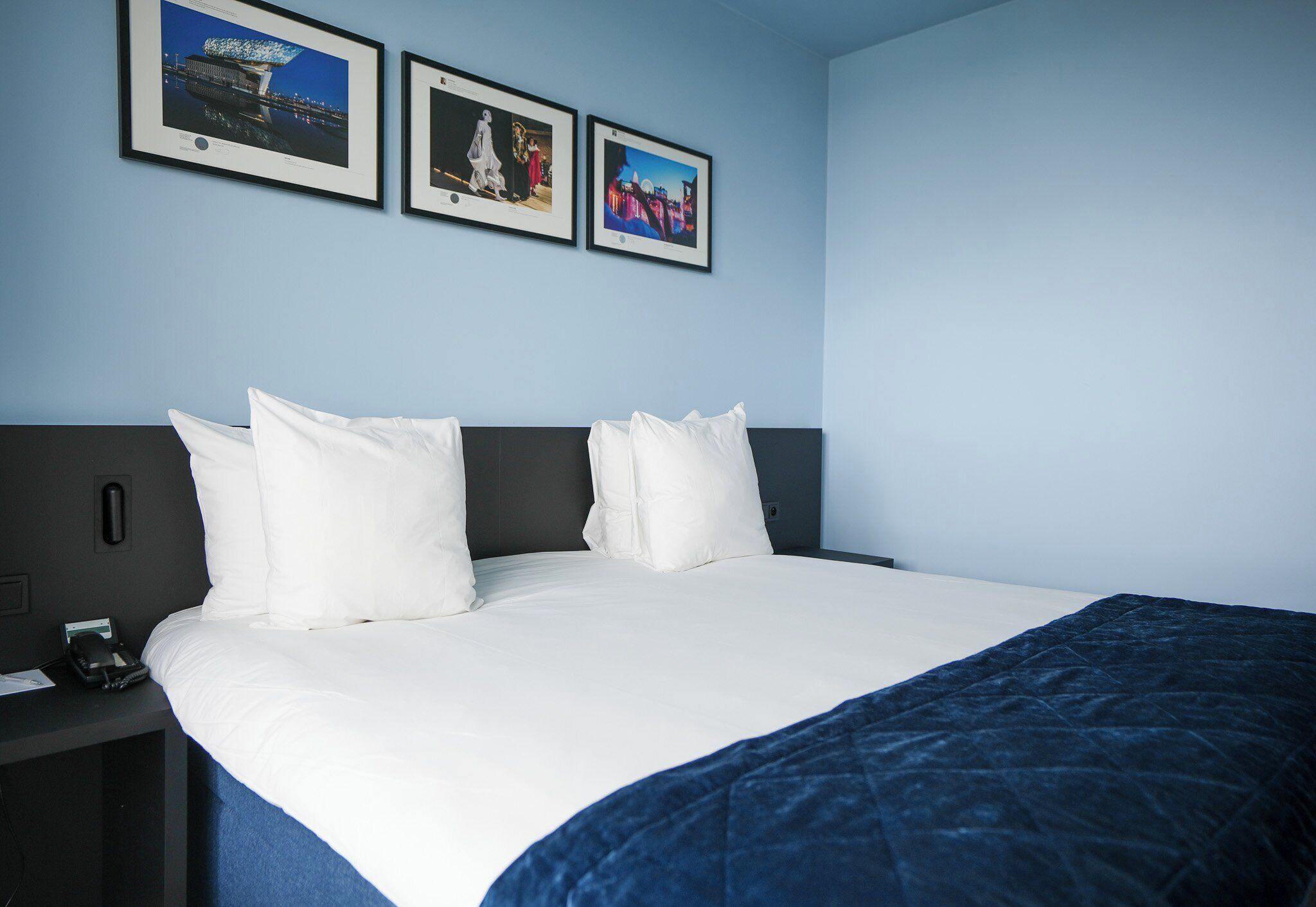 A neatly made bed with white linens and a blue blanket, a bedside table with a phone and papers, and three framed pictures on the wall.