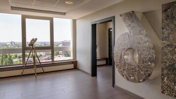 Una habitación amplia con ventanas grandes que ofrecen vista a la ciudad, un trípode con una cámara y una original pieza de arte abstracto en la pared.