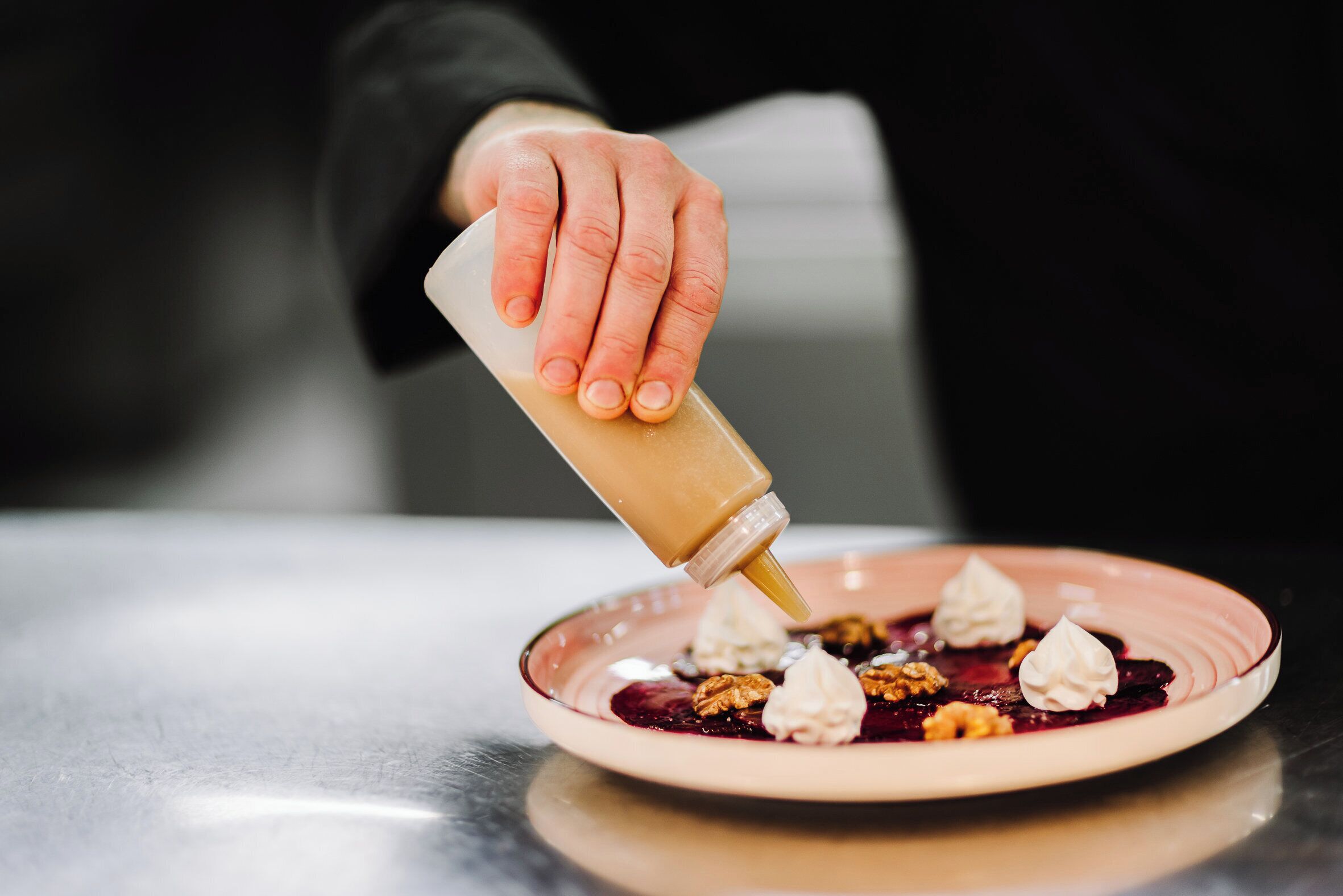A hand pouring sauce onto a dessert plate with whipped cream and fruit.