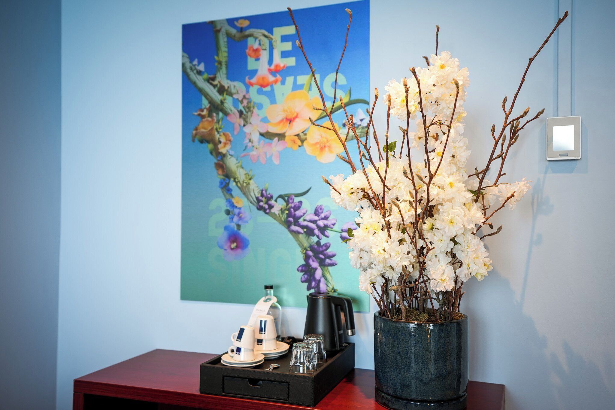 A desk with a coffee maker, cups, and a vase of flowers against a wall with a floral painting.