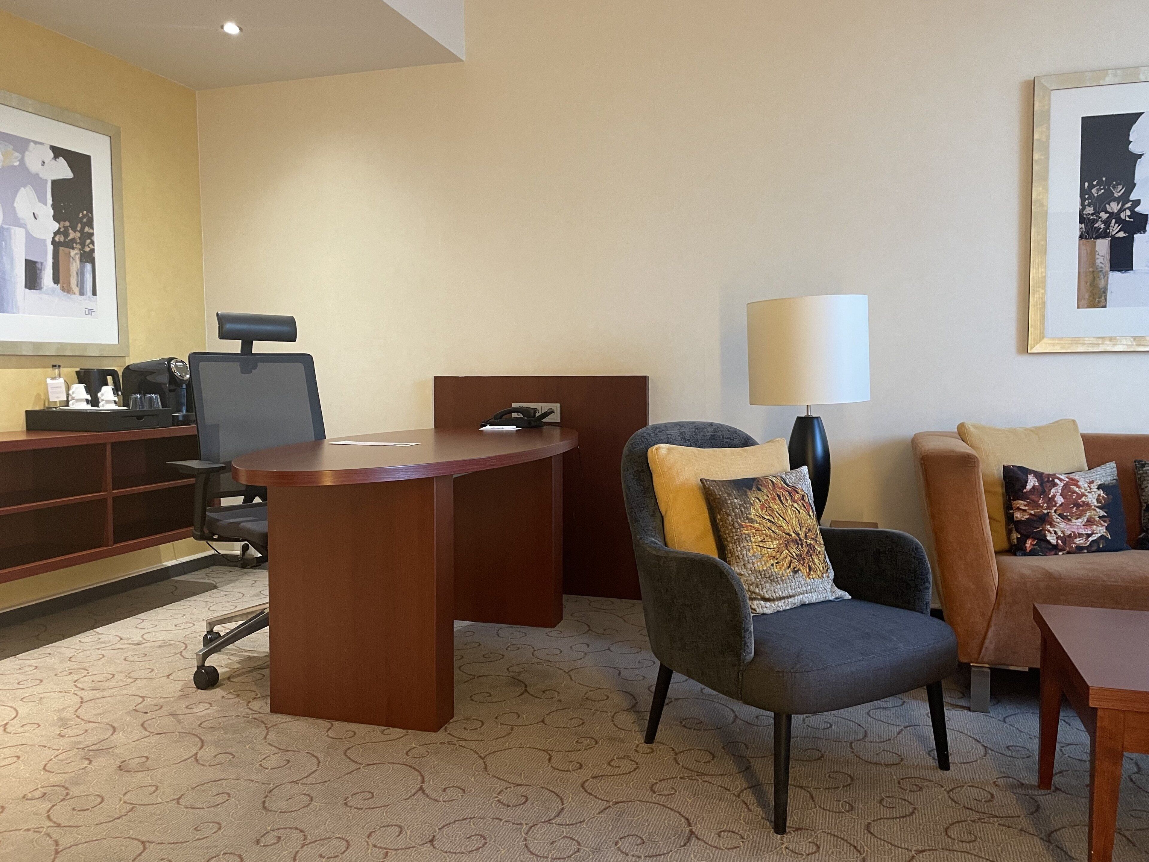A hotel room with a wooden desk, a grey armchair with a patterned cushion, a sofa with decorative pillows, a standing lamp, and a framed artwork on the wall.