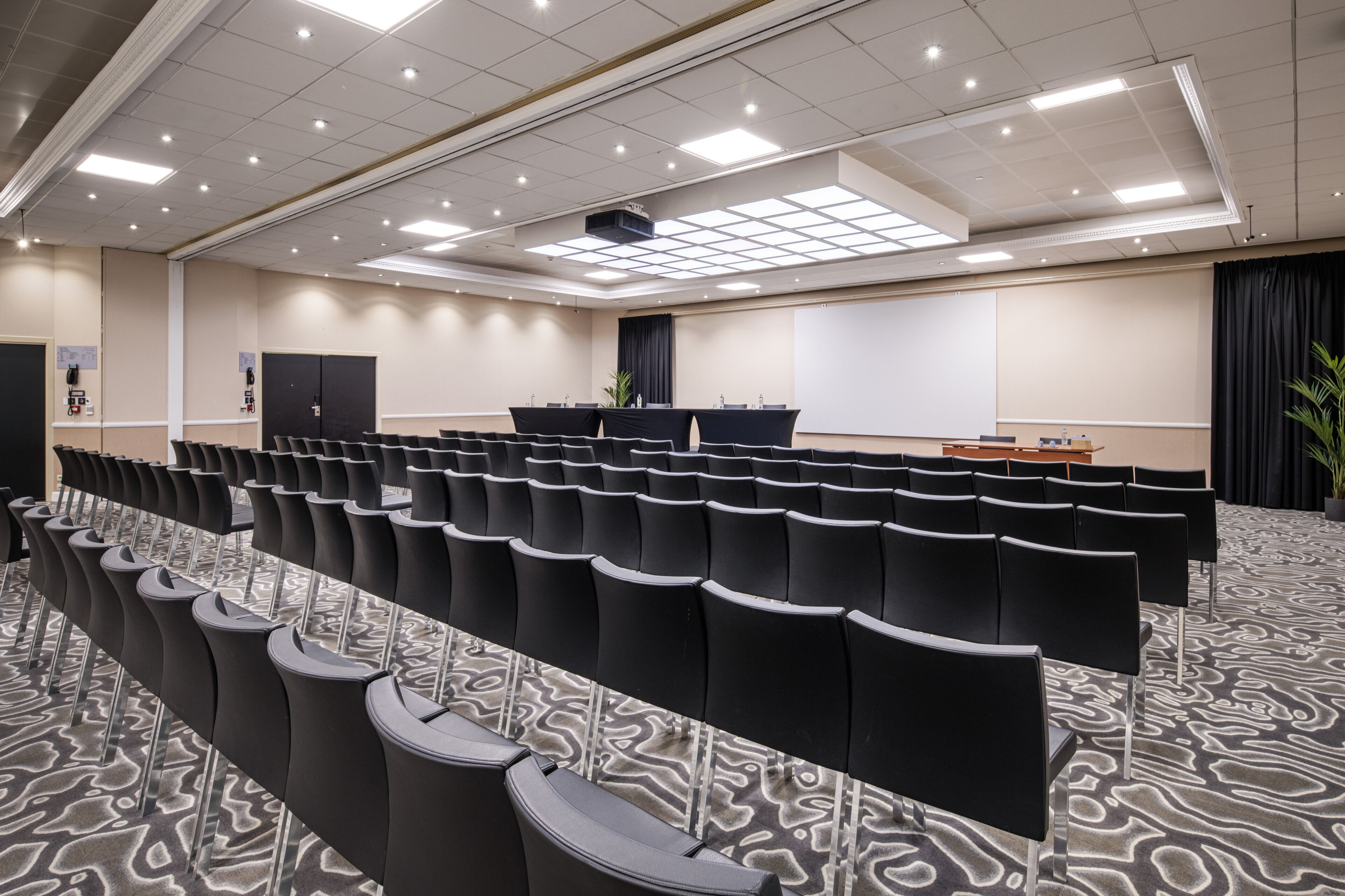 Una sala conferenze con file di sedie nere, uno schermo per proiezioni e un tavolo per presentazioni.