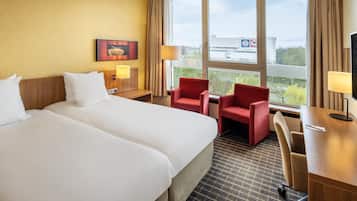Habitación de hotel con dos camas, un escritorio, una silla y un ventanal con vistas a la ciudad.