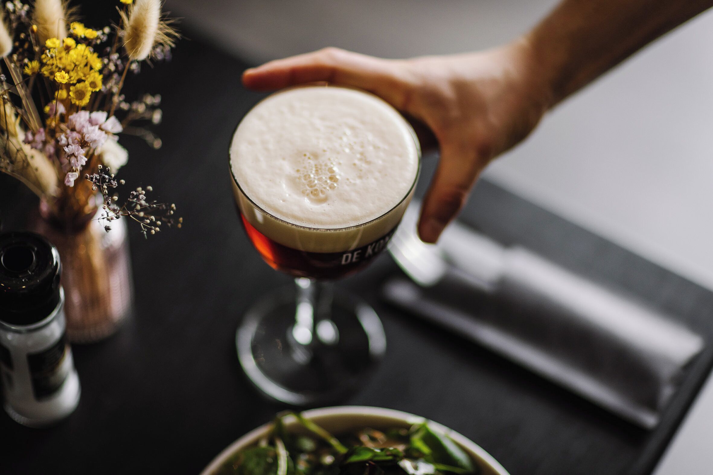 Eine Hand hält ein Glas Bier mit Schaumkrone, Salz- und Pfefferstreuer sowie eine Schüssel mit Salat auf einem Tisch.
