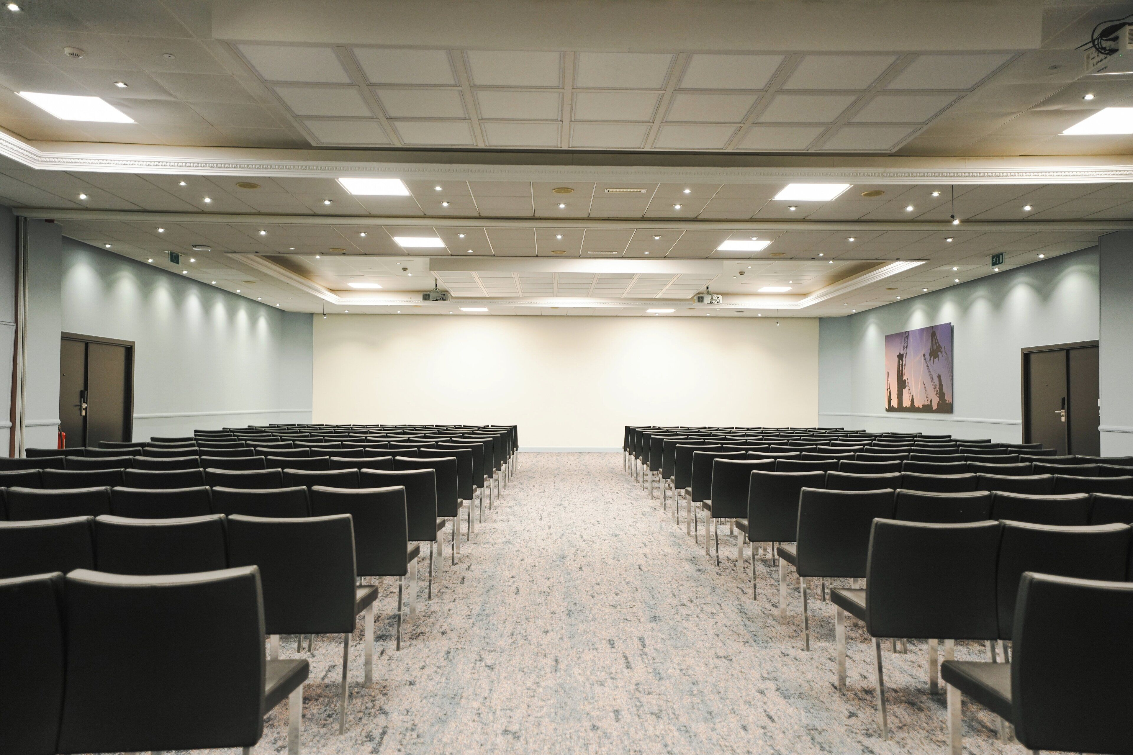 Une salle de conférence avec des rangées de chaises noires, un sol recouvert de moquette et un plafond blanc doté d’un éclairage encastré.