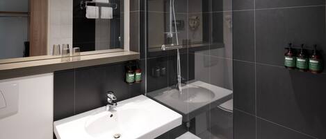 Un baño moderno con lavamanos blanco, cabina de ducha con mampara de vidrio y paredes de baldosas oscuras.