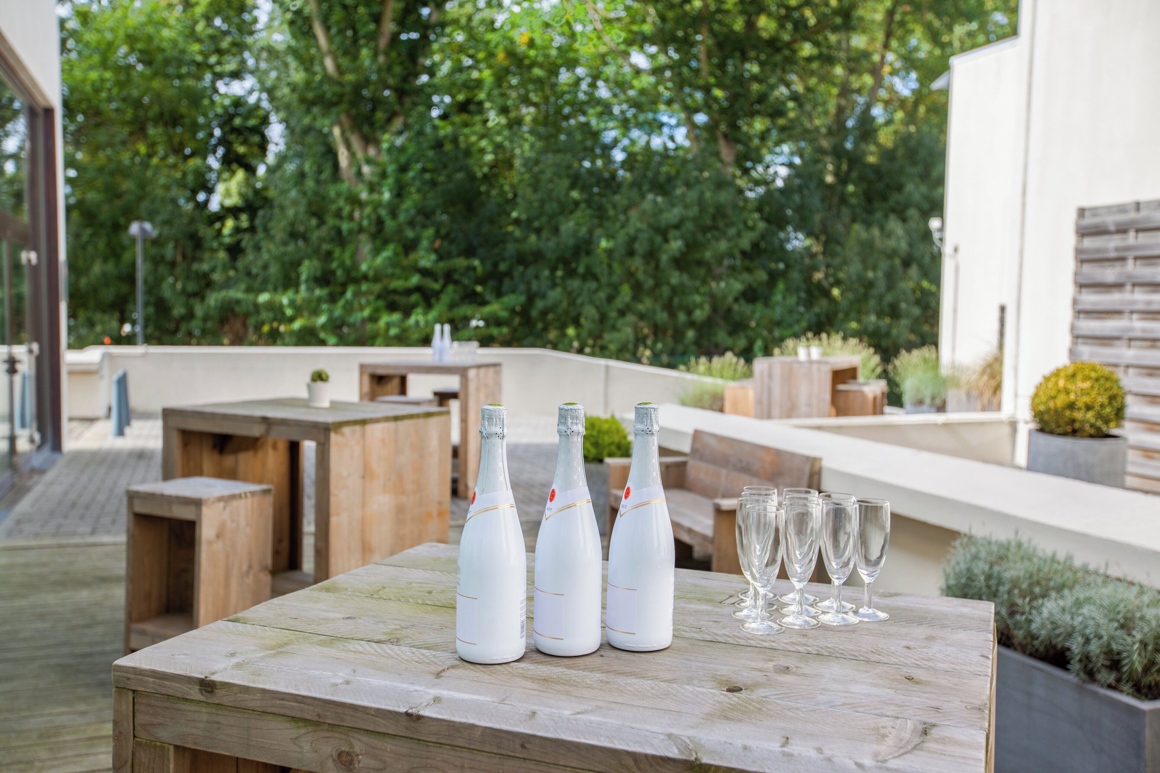 Une table en bois, sur laquelle sont disposées trois bouteilles et six verres, est placée à l’extérieur, offrant une vue sur la verdure et un bâtiment en arrière-plan.
