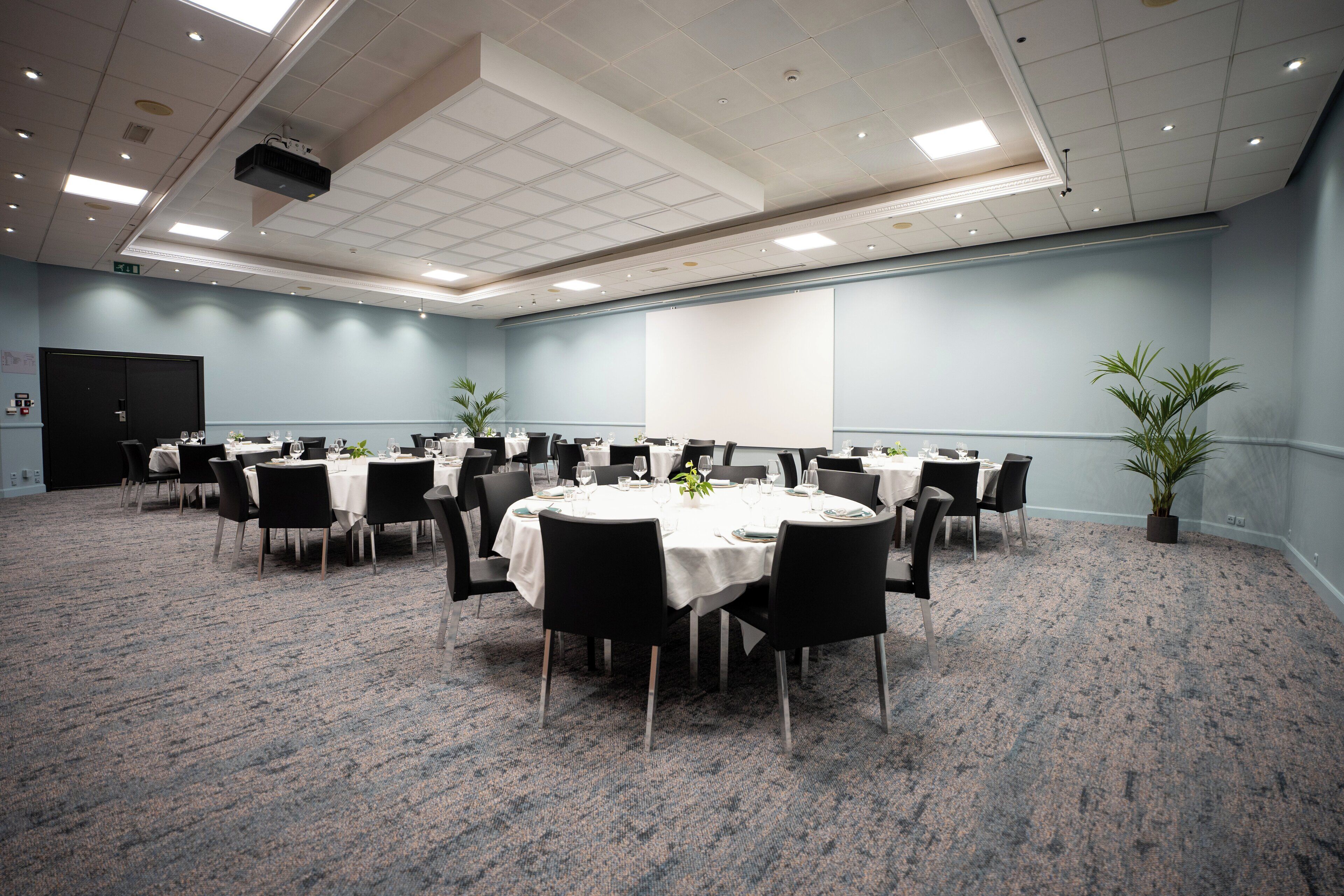 Un salón de conferencias con varias mesas y sillas, un proyector y una pantalla.
