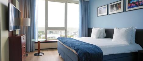 Ein Hotelzimmer mit einem großen Bett, einem Fernseher, einem Schreibtisch mit Lampe und einem Fenster mit Vorhängen.