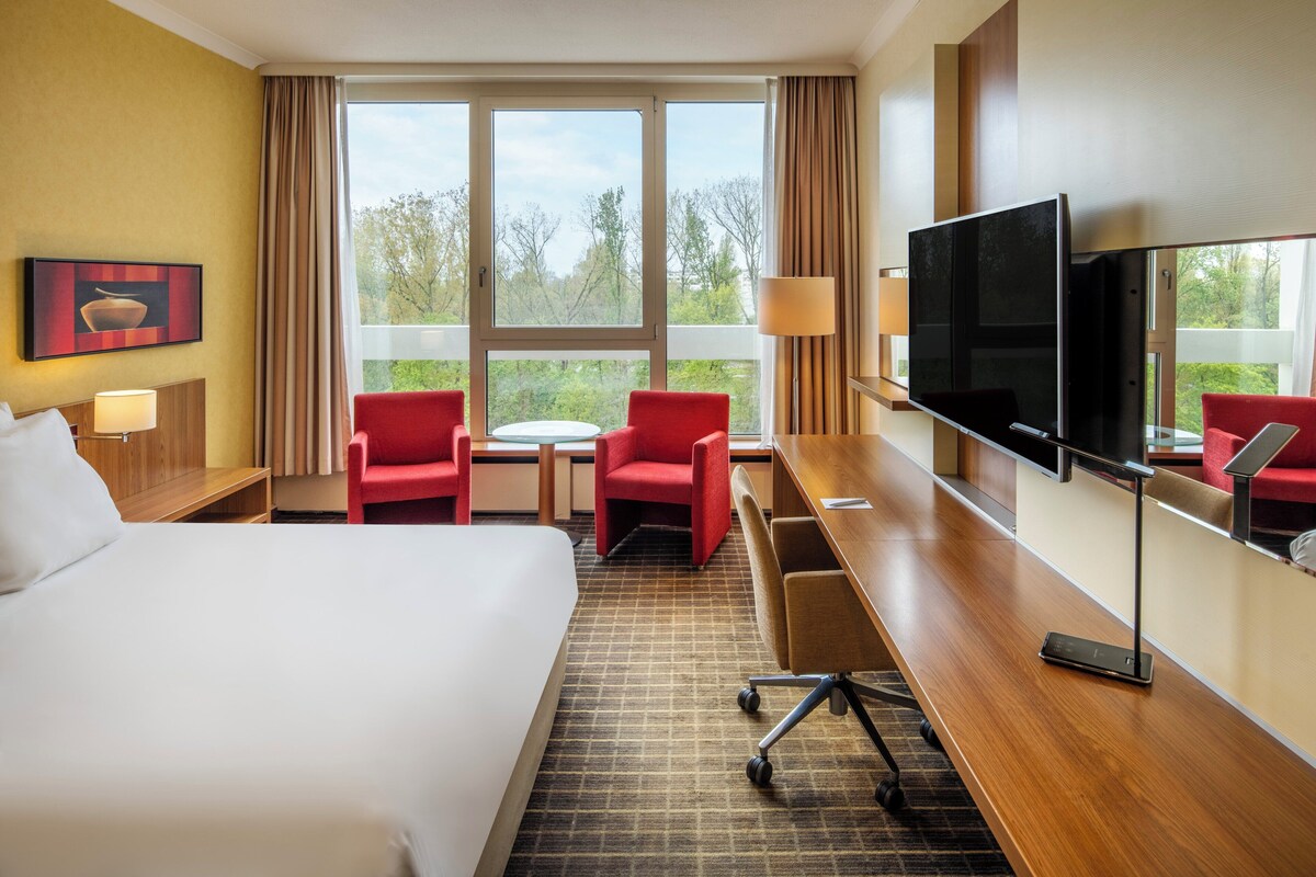 A hotel room with a large bed, two red chairs, a desk with a TV, and a window with a view of trees.