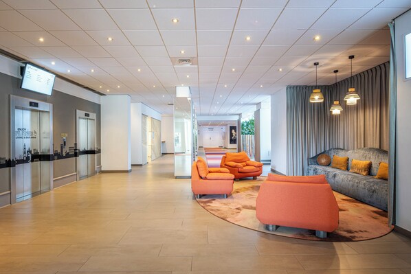 A modern lobby with a reception desk, elevators, orange seating, and a grey sofa with yellow cushions. - Crowne Plaza Antwerp by IHG (Antwerp)