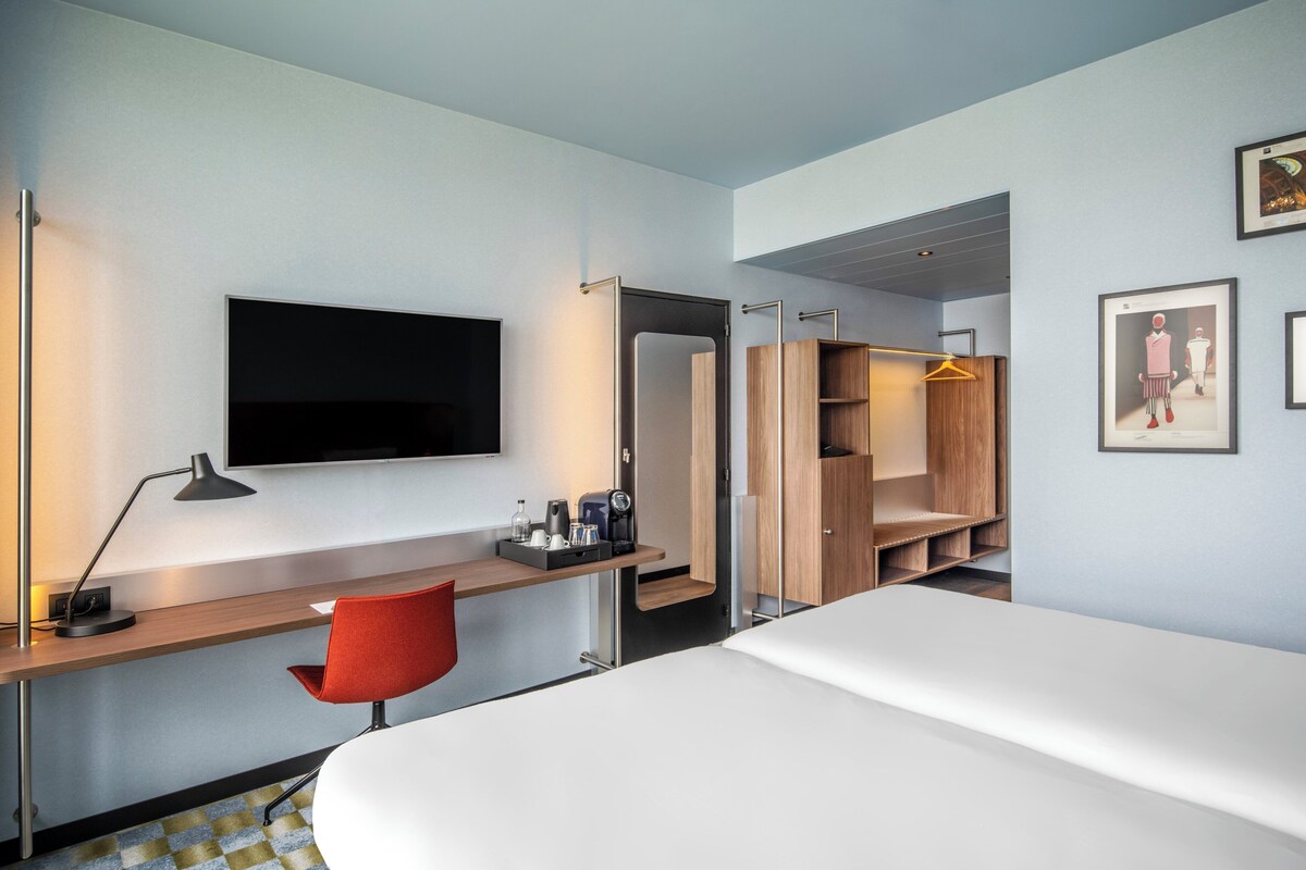 A modern hotel room with a bed, desk, chair, TV, wardrobe, and a framed picture on the wall.