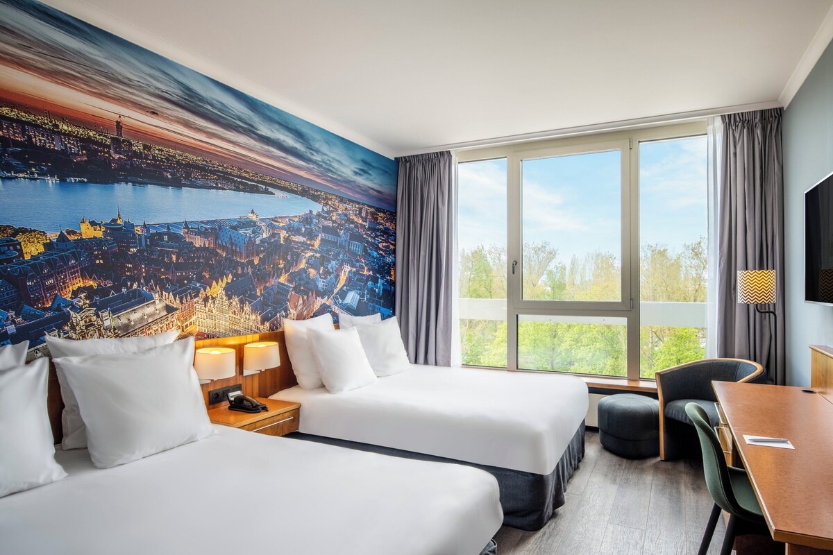 A hotel room with two beds, a large window with a view, a dining table, and a TV.