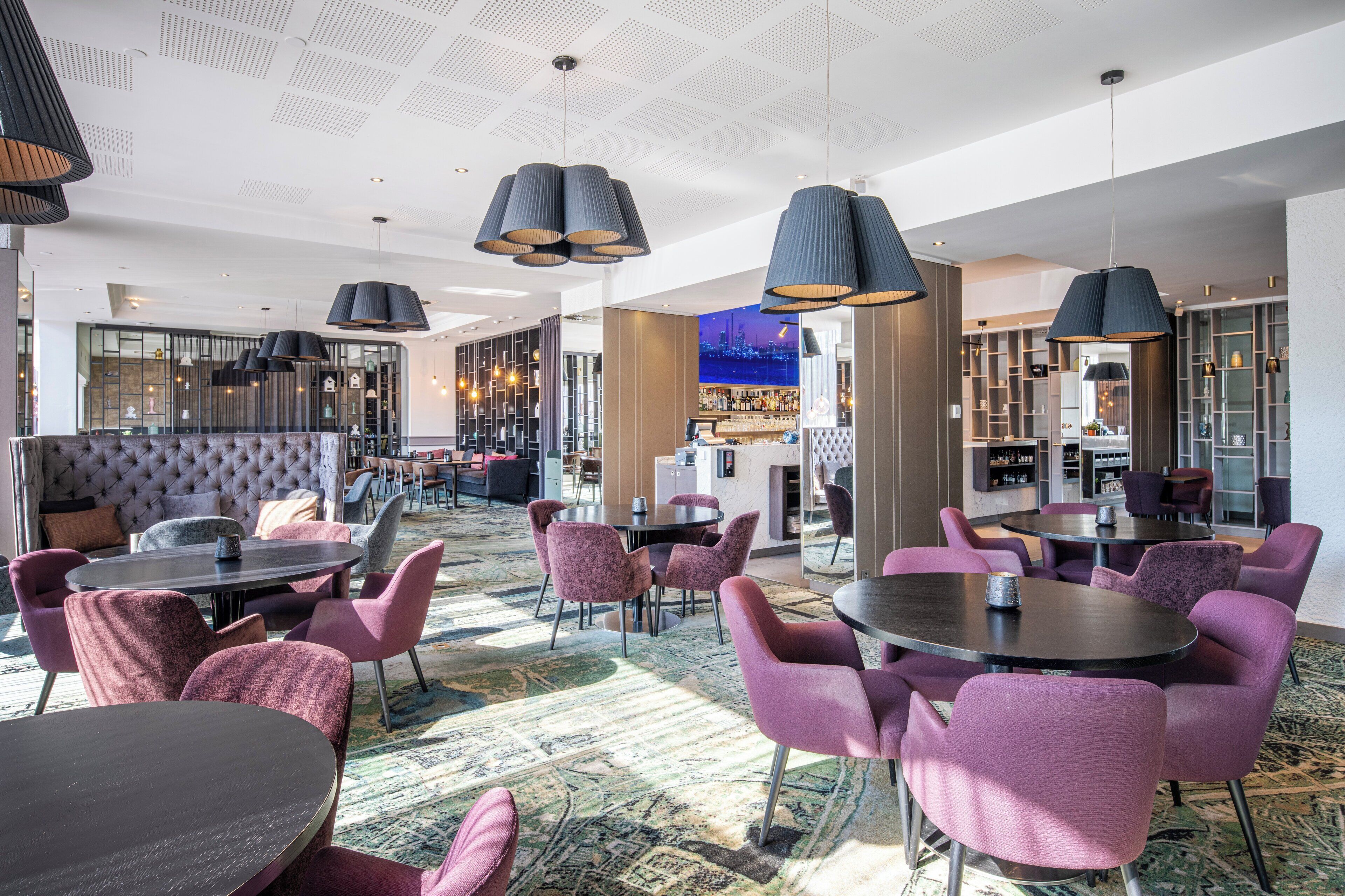 A hotel lobby with round tables and purple chairs, a bar area, and a seating area with a tufted sofa.