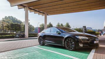 A black electric vehicle charging at a station with a green parking space, a covered area, and a traffic light in the background.
