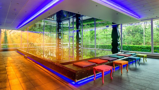 A modern indoor swimming pool with clear glass walls, a wooden deck, and colorful seating. - Crowne Plaza Antwerp by IHG (Antwerp)