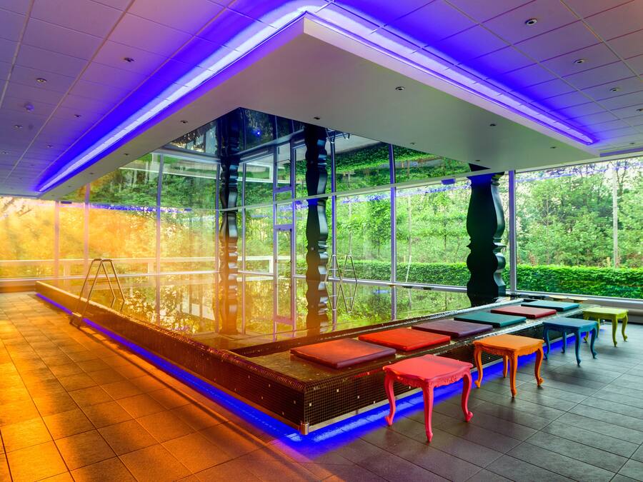 Una piscina interna moderna con pareti di vetro trasparenti, un solarium in legno e sedute colorate.