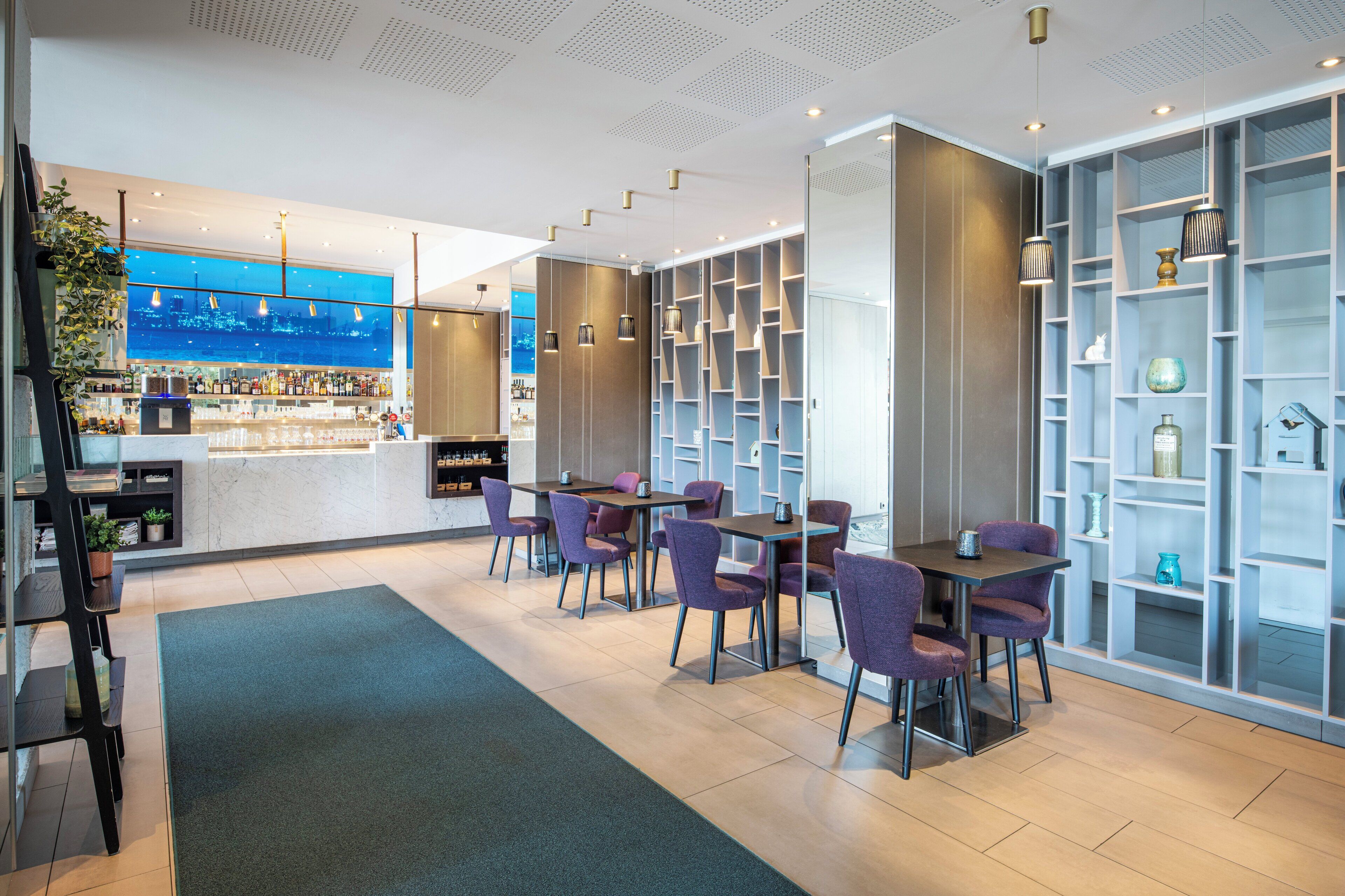 Un hall d’hôtel moderne doté d’un bar, d’espaces aménagés avec des sièges et d’étagères décoratives.