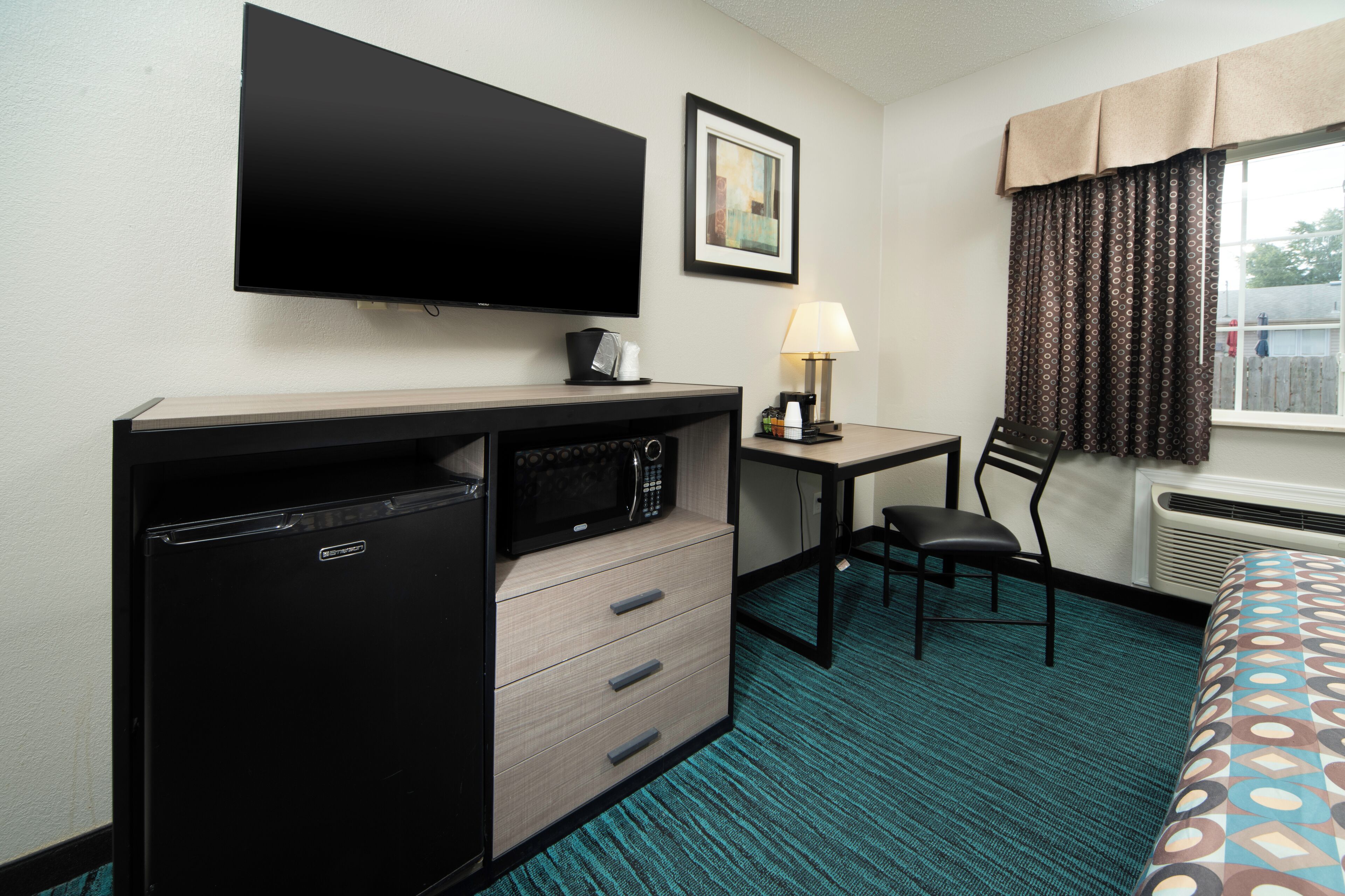 A compact hotel room with a bed, a desk with a chair, a microwave, a mini fridge, a TV, and a window with curtains.