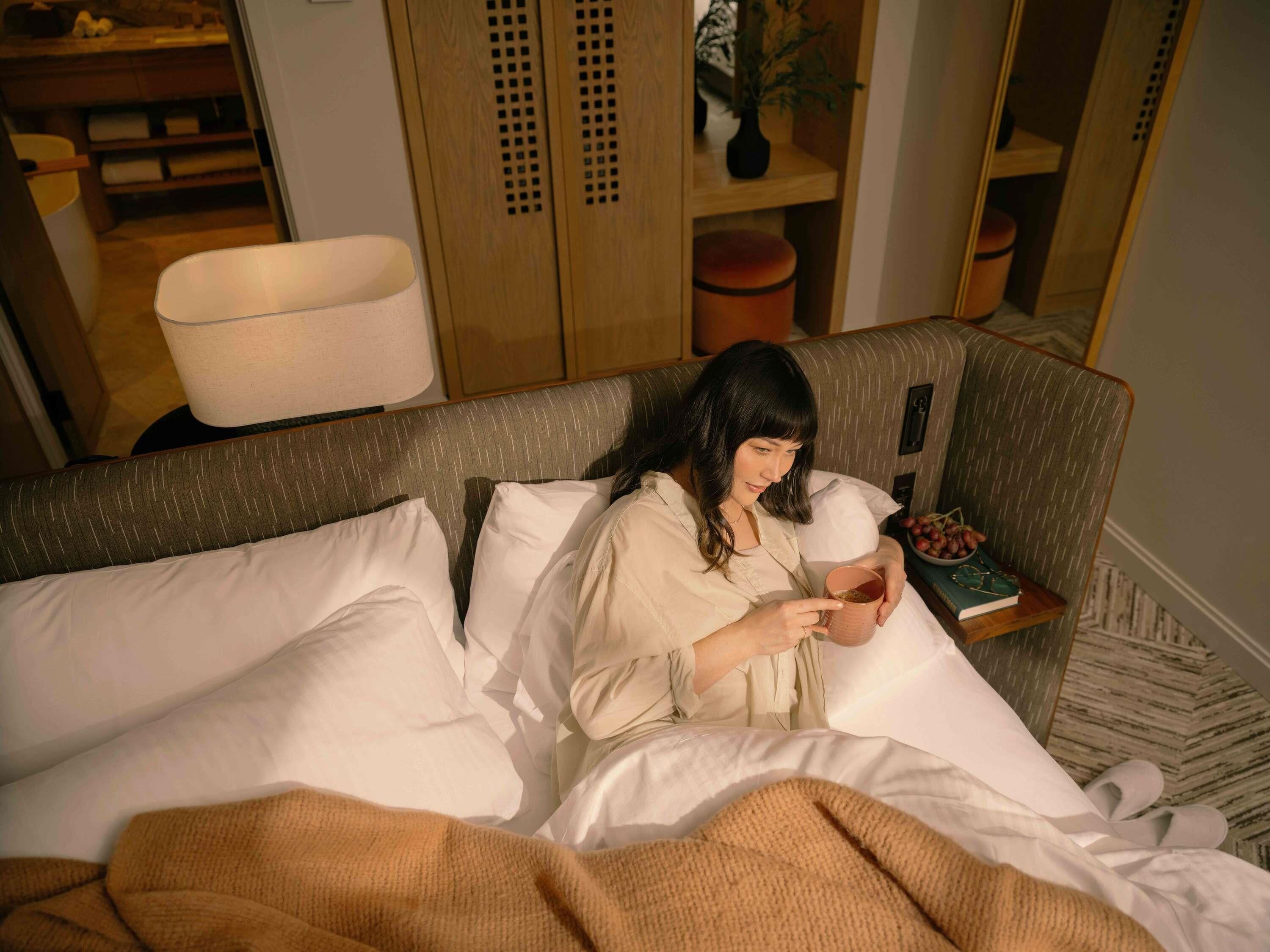 A person lying in bed with a book and a cup, in a room with a wooden wardrobe and a mirror.