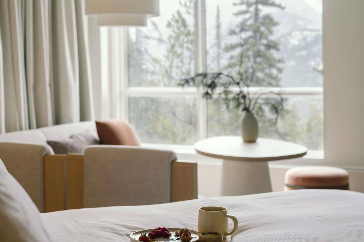 Habitación de hotel con cama, mesita de noche con una taza y frutos rojos, una silla y vistas a árboles por la ventana.