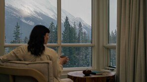 A person sitting by a window with a view of snow-covered mountains and trees. - Rimrock Banff, Emblems Collection (Banff)